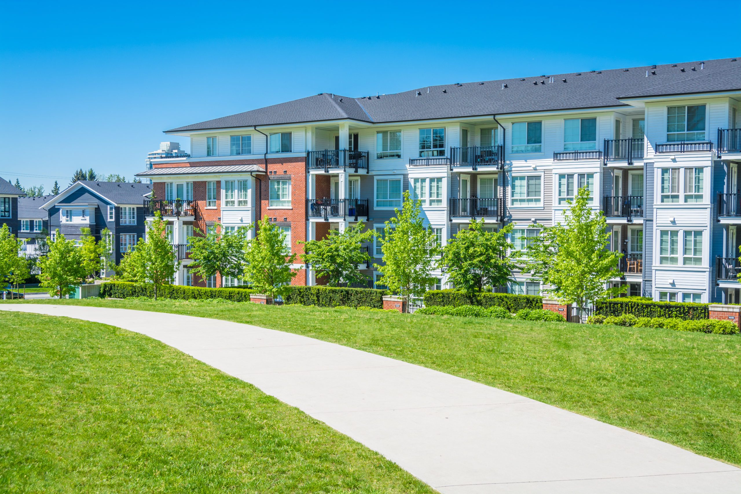 Concrete pathway across green lawn in front of residential condo building. Residential apartment building on sunny day with blue sky.